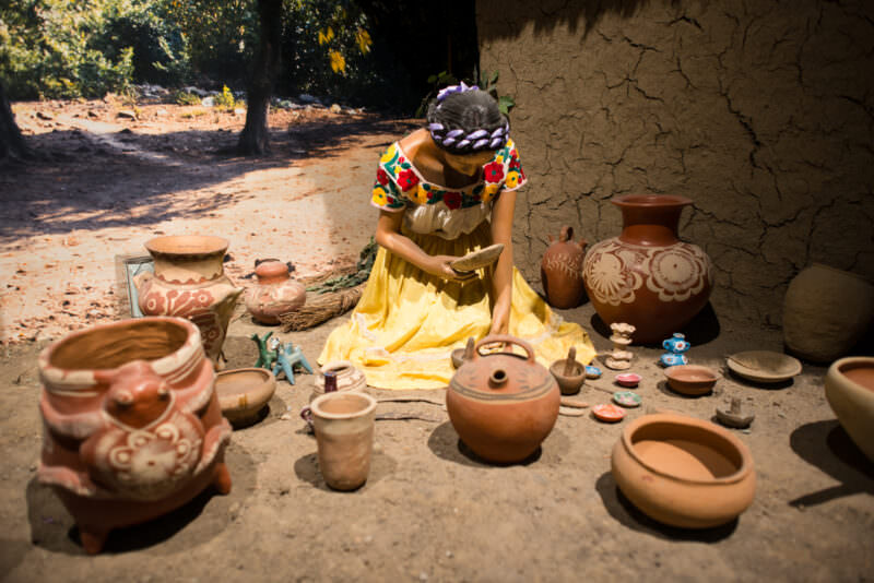 Life-size diorama of woman in traditional dress finishing ceramic vessels by hand beside adobe wall, varied pottery around her