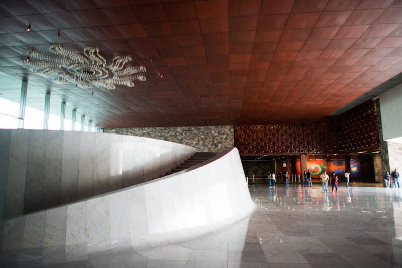 Museo Nacional de Antropología lobby with sweeping marble staircase, tezontle ceiling, and aluminum serpent-motif celosía on far wall