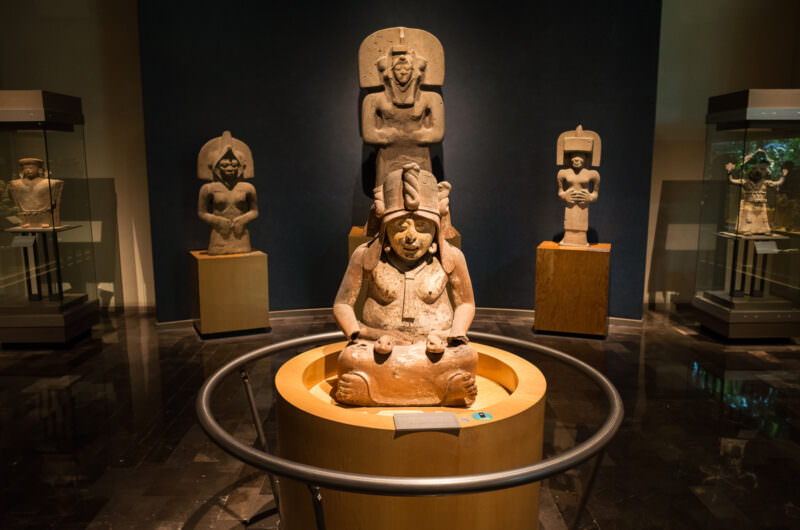 Seated ceramic deity figure with tall headdress in circular display, flanked by standing stone sculptures
