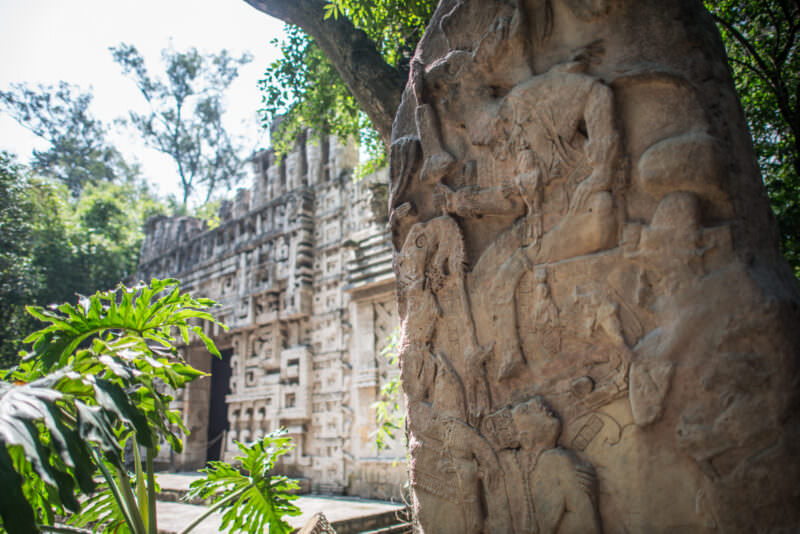Carved stone stela with figural Maya relief in foreground, Puuc-style palace facade replica rising through trees behind