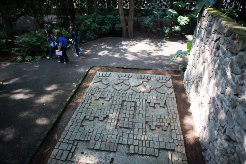 Stone mosaic floor panel in outdoor garden rendering stylized Mesoamerican mask face in fitted tesserae beside rubblework wall