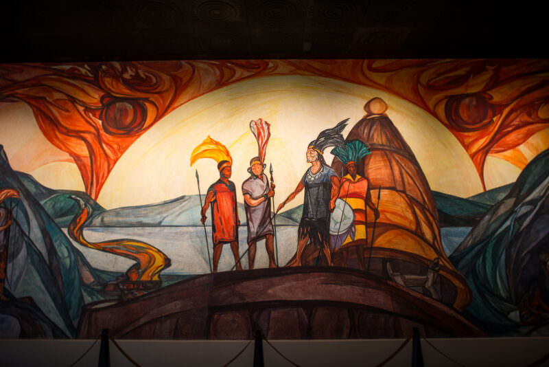 Mural depicting armed warriors with feathered headdresses and spears beneath swirling flames and luminous sky arch