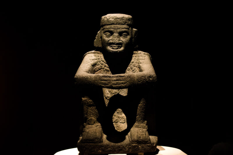 Stone figure seated with arms folded across chest and flat headdress, against dark background