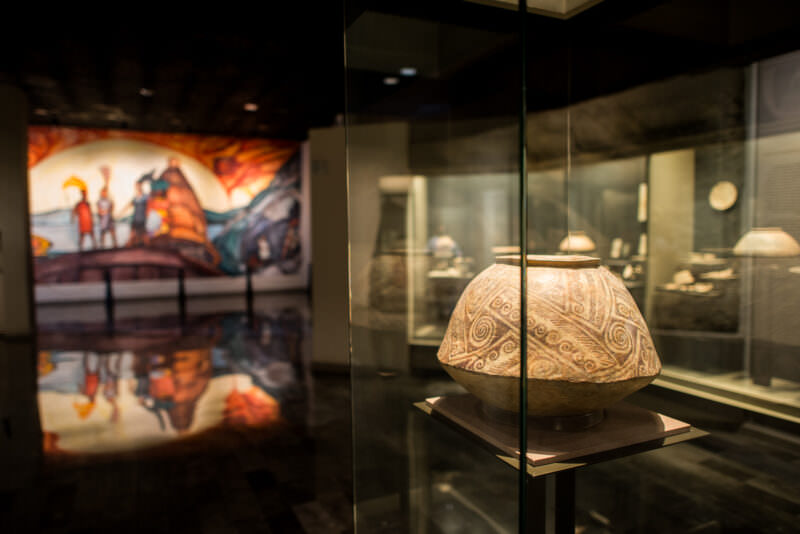 Large incised ceramic vessel in glass case with vivid mural backdrop depicting Mesoamerican life in gallery behind