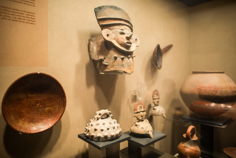 Ceramic effigy head mounted high on wall above mixed display of vessels, figurines, and painted bowl in regional hall