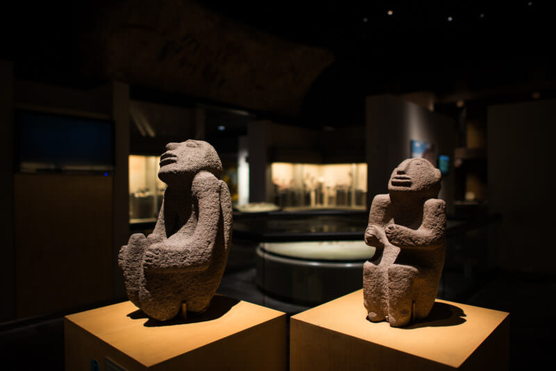 Two rough-hewn stone figures, wide-shouldered with arms folded across chest, in foreground of archaeology gallery