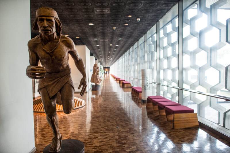 Bronze running figure in upper corridor beside geometric aluminum lattice screen filtering afternoon light
