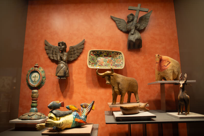 Carved wooden angels, animal figures, painted ceramic tray, and monstrance grouped in ethnography display illustrating Catholic syncretism