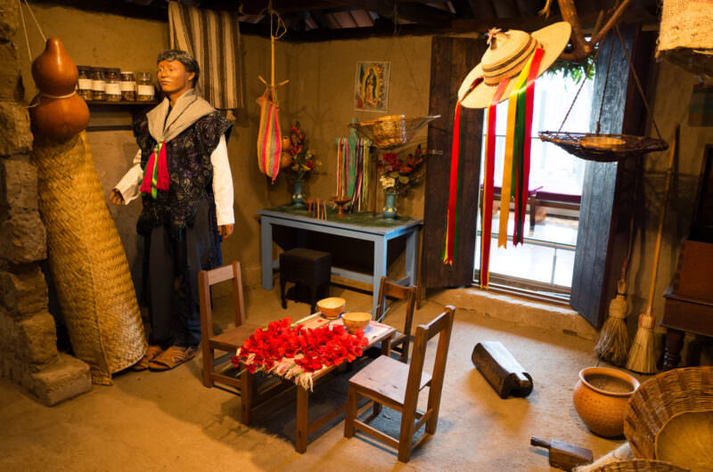 Ethnographic diorama with male figure in serape beside home altar bearing Virgin of Guadalupe image, candles, and red flowers