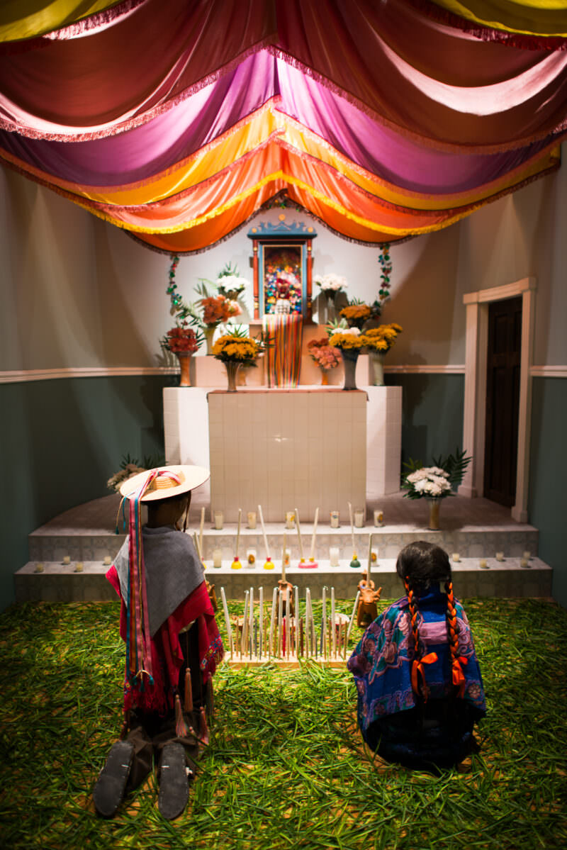Two mannequins in traditional dress kneeling before flower-decked church altar on bed of fresh grass in ethnographic diorama