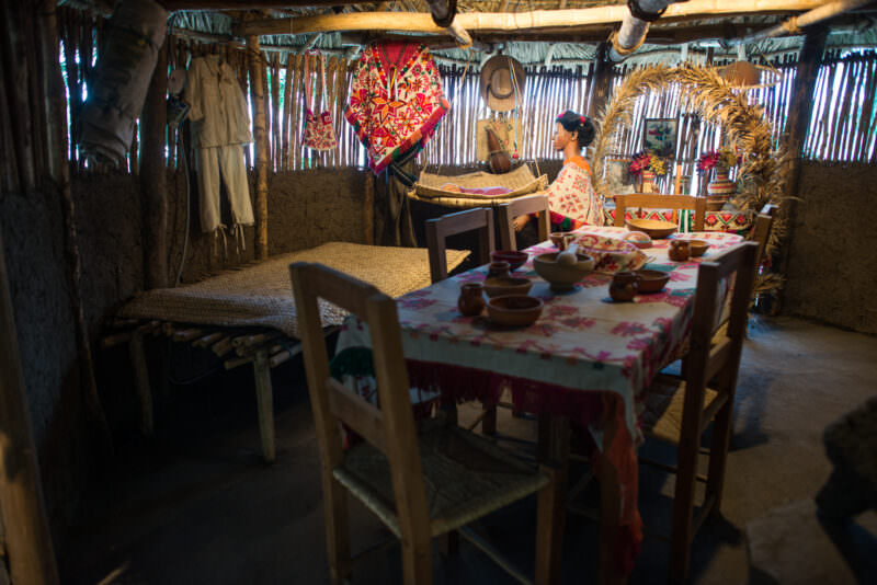 Life-size ethnographic diorama of rural indigenous dwelling interior with mannequin in embroidered dress, clay bowls, woven mat, textiles, and basketry