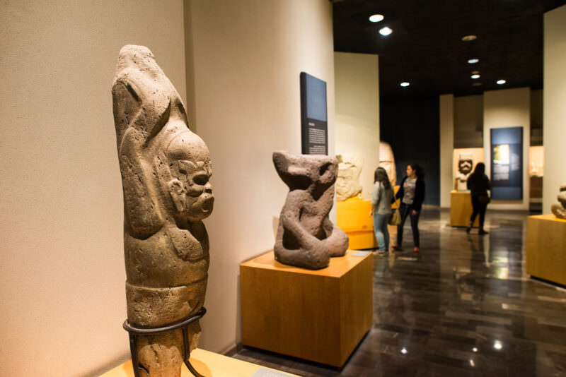 Visitors examining stone sculptures, tall carved figure in foreground and compact seated figure on pedestal anchoring display