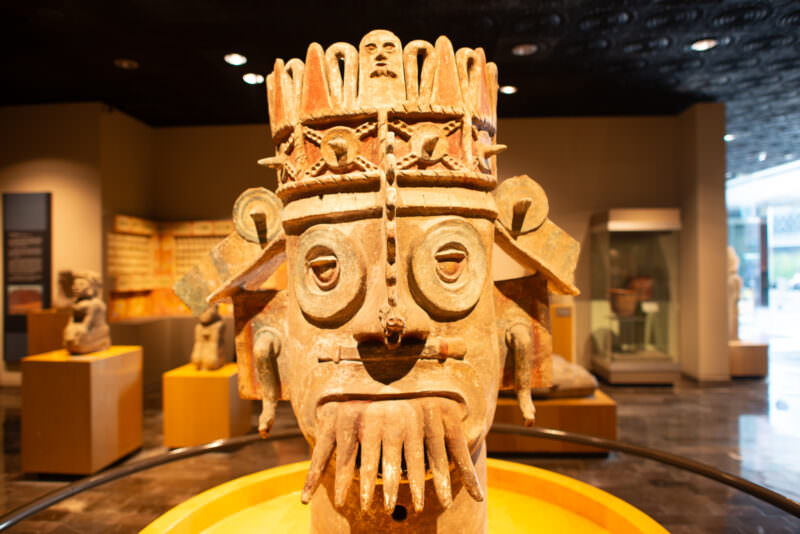 Large polychrome ceramic deity head with goggle eyes, fanged mouth, elaborate headdress bearing small skull, in center of gallery