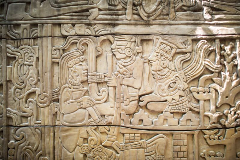 Large carved relief panel showing Maya figures in ritual scene with feathered headdresses, scrollwork, and glyphic borders