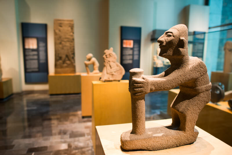 Granular-stone figure of kneeling man gripping vessel in foreground, additional figurative sculptures receding into softly lit room