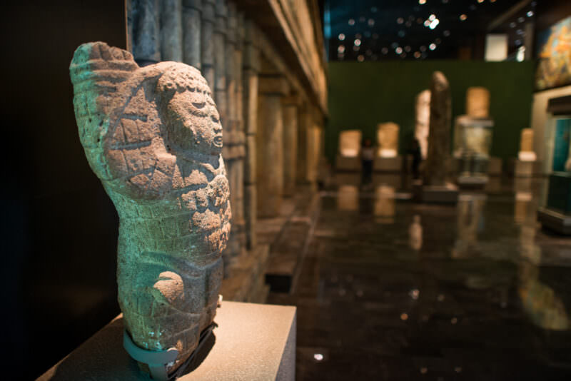 Stone figure with raised arm in ground-floor galleries, dark-red tezontle-clad walls of Mexica hall visible behind