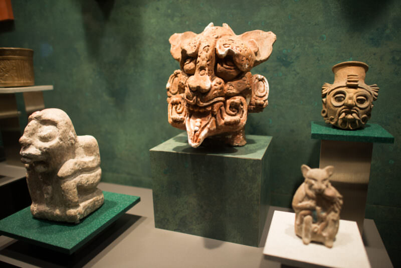 Display case of pre-Columbian ceramic deity figures, vivid terracotta head with fanged open mouth and headdress dominating center