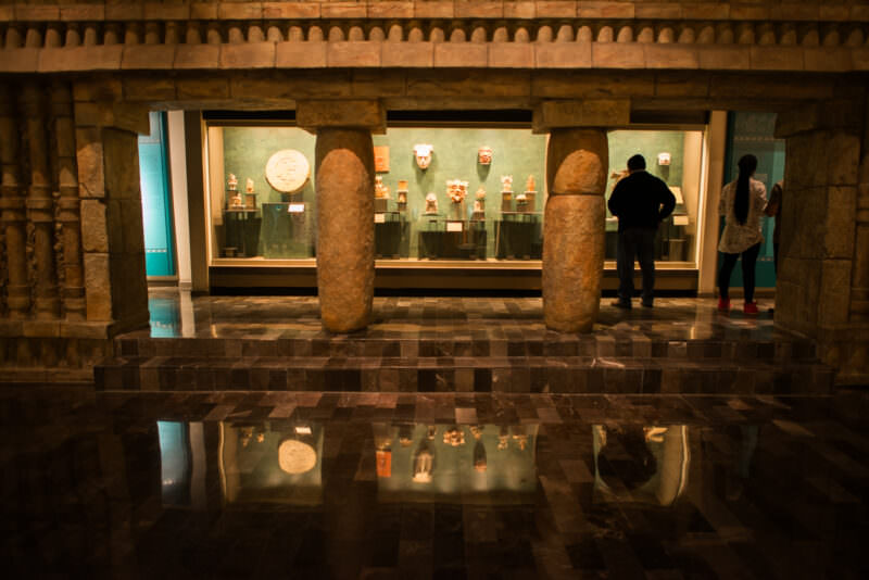 Two visitors examining backlit display case of Maya ceramics and masks framed by cylindrical stone columns, reflections on polished marble floor