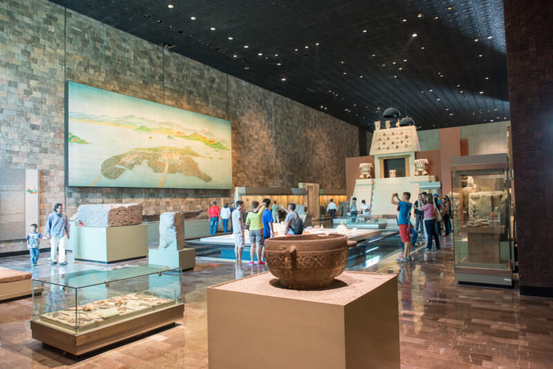Mexica hall with visitors viewing mural of Tenochtitlan, carved stone vessel in foreground, dark volcanic tezontle walls