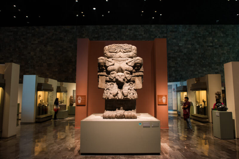 Monumental Coatlicue basalt statue with severed-serpent-head collar, skull belt, and clawed hands in center of Mexica hall