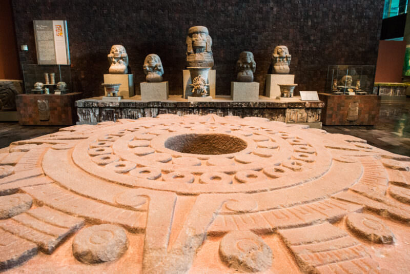Circular stone altar with hollow central depression and relief bands, row of deity busts on platform behind, dark tezontle walls