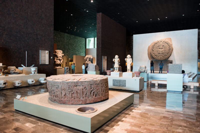 Mexica hall with Piedra del Sol on rear wall, large cylindrical sacrificial stone carved with processional figures in foreground