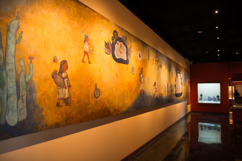 Large mural depicting indigenous figures carrying goods and shields under swirling flames and luminous sky