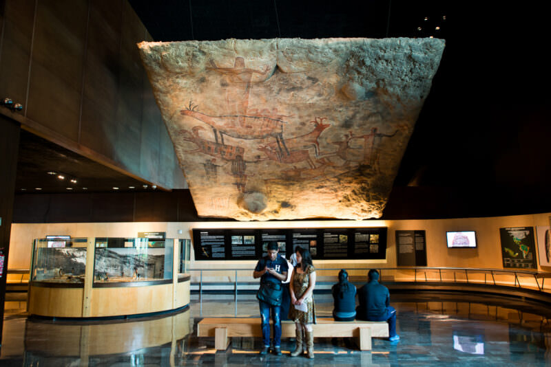 Large-scale rock art replica suspended overhead like a cave ceiling, painted deer, human figures, and abstract forms visible from below