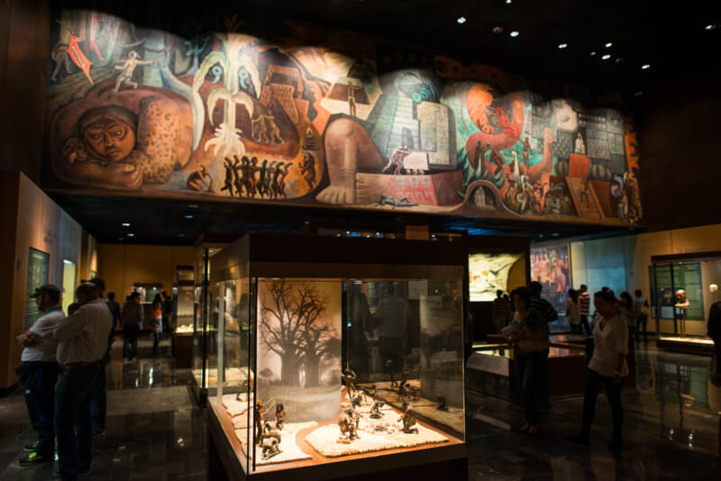Sweeping mural depicting pre-Columbian civilization arc spanning upper wall of introductory hall, visitors below examining display cases