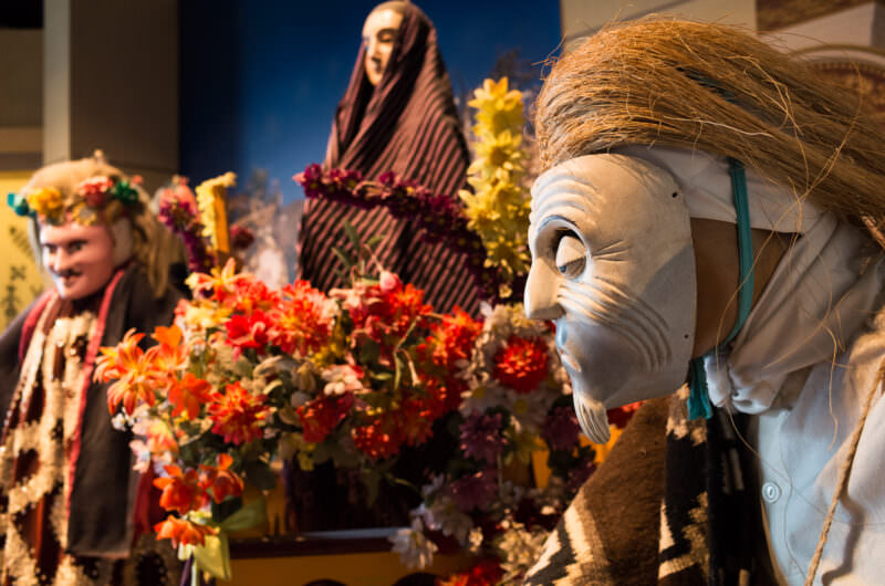 Masked and costumed figures with paper flowers in Día de Muertos diorama, pale wrinkled mask in foreground
