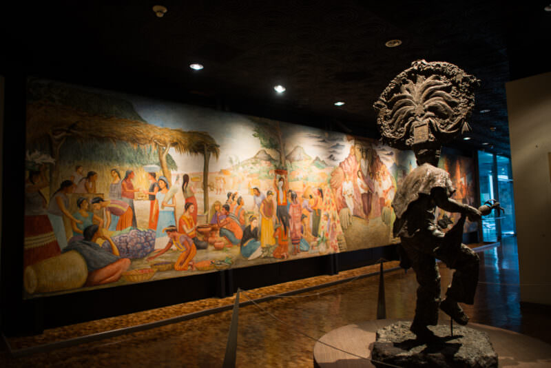 Large ethnographic mural of indigenous market scene in Oaxaca hall paired with bronze sculpture of figure bearing ceremonial headdress