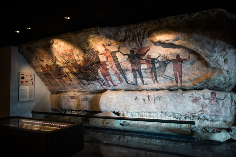 Full-scale rock art replica showing red, black, and yellow human figures and deer painted on cave-surface mock-up