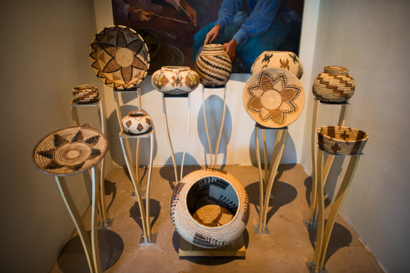 Seri coiled baskets and vessels from Sonora on individual stands, painting of weaver visible behind