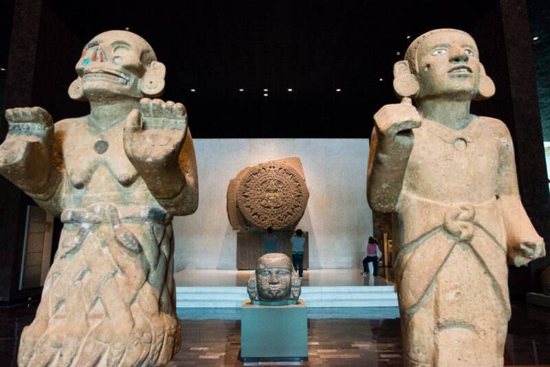 Two monumental Mexica stone figures flanking the Piedra del Sol, with severed deity head on plinth between them