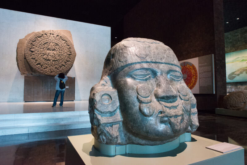 Visitor studying the Piedra del Sol on wall, colossal carved stone head with blue-green pigment in foreground