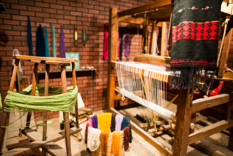 Green wool skein on warping frame beside loaded treadle loom, black-and-red geometric textile draped across loom beam