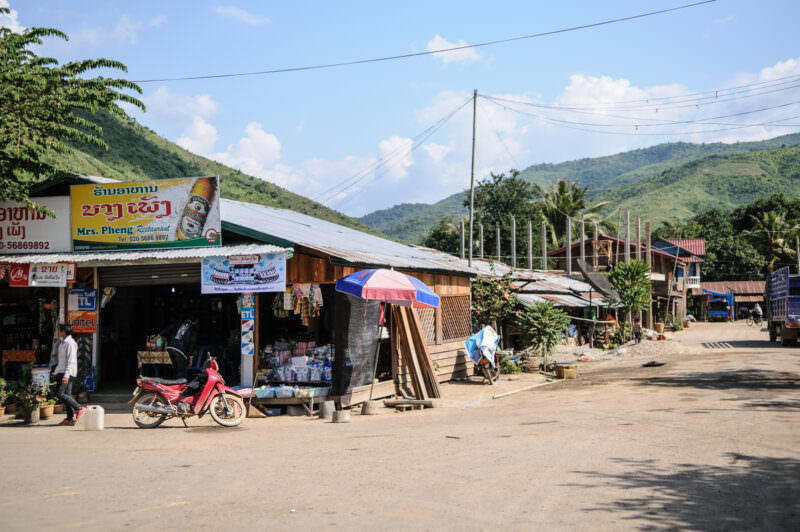 Mrs. Pheng Restaurant wooden shopfront with Beerlao signage, red motorbike parked in front, mountains and dirt road visible