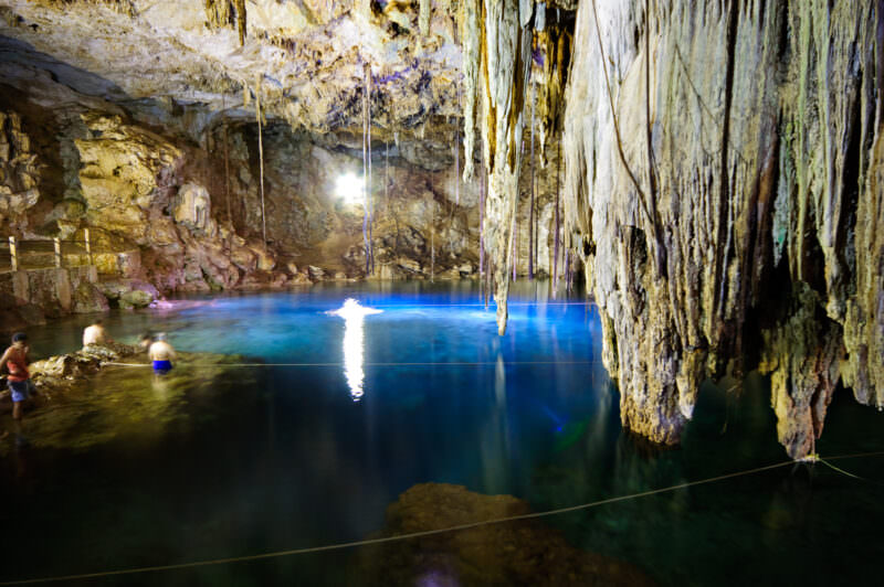 Long limestone stalactites hanging from Cenote X'keken's ceiling, illuminated by natural light from above, with swimmers visible in the turquoise water below