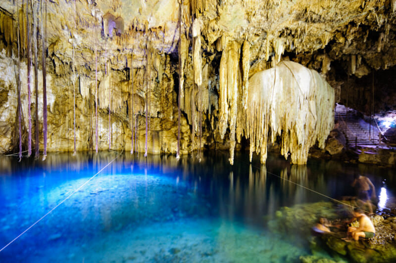 Cenote X'keken's underground pool with dramatic floor-to-ceiling stalactite columns and a curtain of hanging formations, lit by natural and artificial light