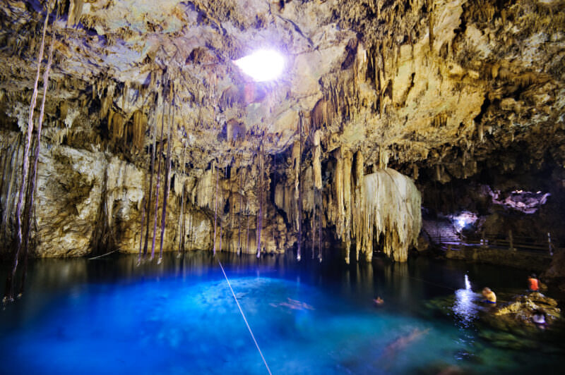 Cenote X'keken's illuminated turquoise pool beneath a ceiling opening that casts natural light, surrounded by extensive stalactite formations and viewing platforms