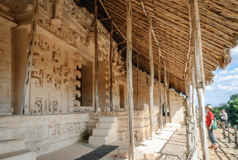 Visitor photographing the stucco façade of the Sak Xok Naah on Ek' Balam's Acropolis under a thatched shelter