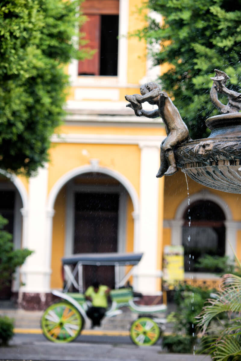 Bronze cherub holding a shell spraying water in the central fountain of Parque Central, yellow colonial building behind