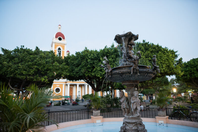 Cherub fountain in foreground with yellow neoclassical Granada Cathedral and red domes across Parque Central at dusk
