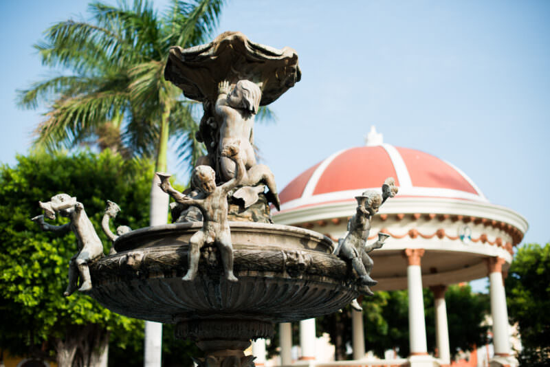 Stone cherubs on classical fountain with red-domed gazebo behind palm trees, Parque Central Granada