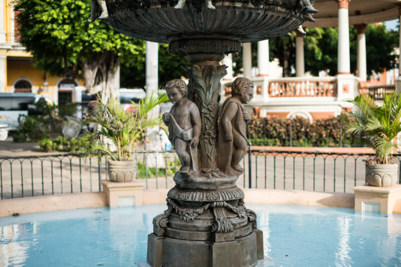 Bronze toga-draped figures on fountain pedestal with red-domed gazebo in background, Parque Central Granada