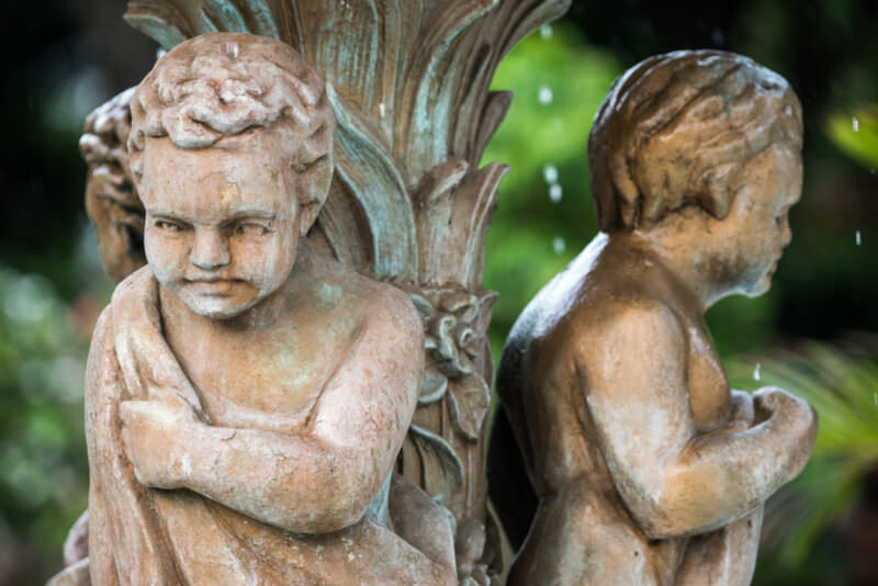 Two weathered fountain sculptures of boys in Roman togas beside bronze acanthus leaves, greenery behind