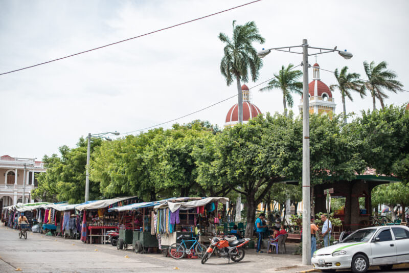 Souvenir stalls beneath trees at Parque Central, red cathedral domes visible above the tree line