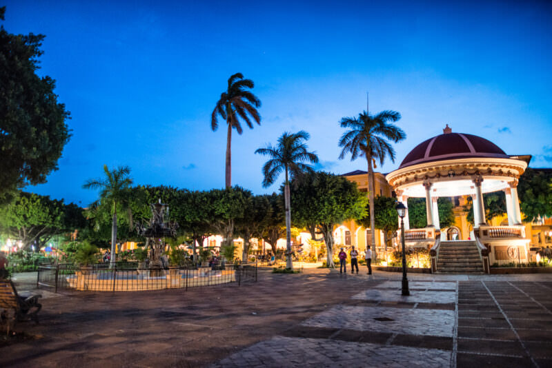 Red-domed bandstand and ornate fountain in Parque Central at dusk, tall palms silhouetted against blue sky