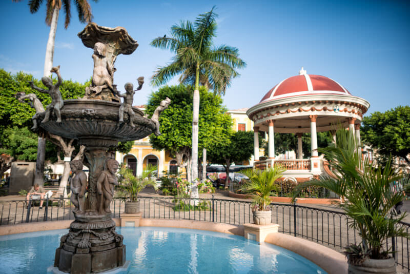 Ornate cherub fountain in Granada's Parque Central with red-domed bandstand visible behind palm trees