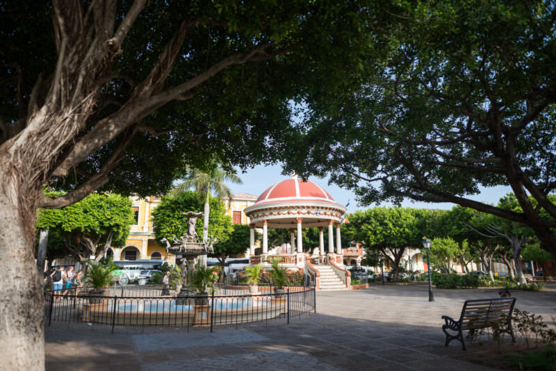 Ornate fountain and red-domed neoclassical kiosk shaded by large trees in Parque Central Granada